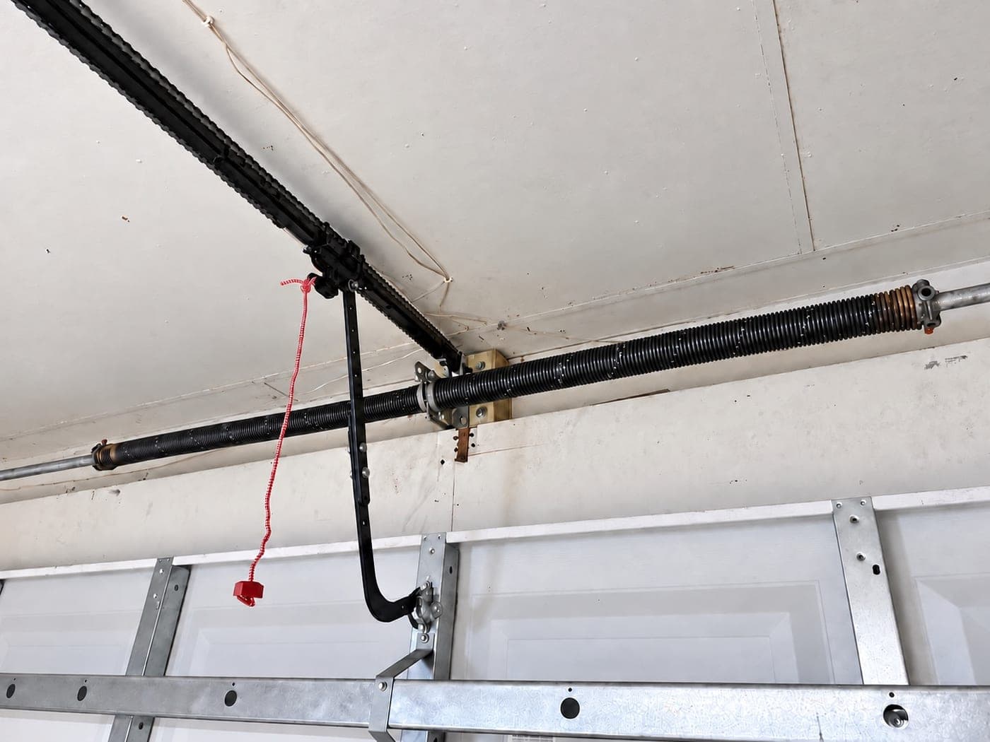 Torsion spring mounted on the shaft above a white sectional garage door, with the opener rail and red emergency release cord